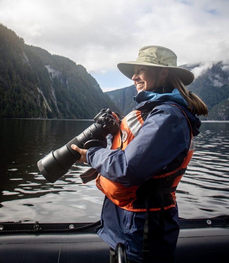 Delaney in her natural habitat Woman in rain gear holding a camera standing in a Zodiac with a fjord in the background