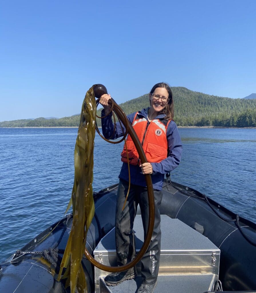 Jenny AKA Science Mom Woman holding a piece of Bull Kelp at the bow of a Zodiac on a cloudless day