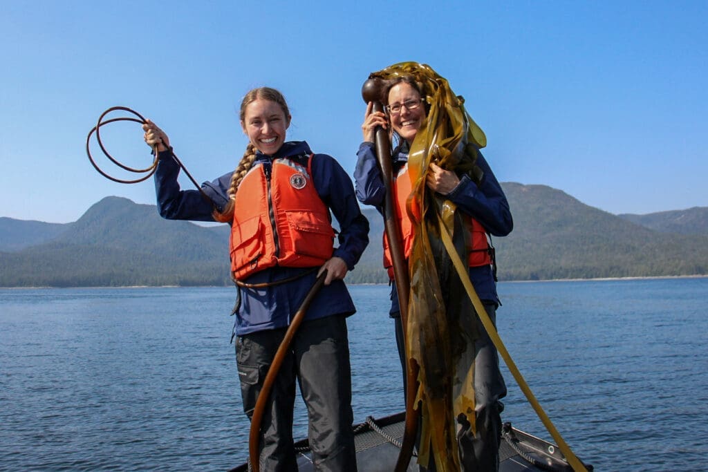 Bull Kelp Ladies Two women in rain gear and life jackets pose on the bow of a Zodiac with a piece of bull kelp wrapped around them